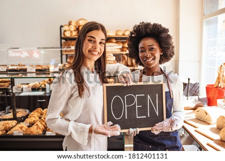 Portrait of two young entrepreneurs standing in the bakery shop with OPEN sign. Two cheerful small business owners smiling and looking at camera while standing with open sign board.