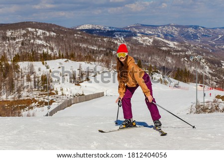 Young beautiful woman in red hat and glasses stands on skis on the mountain in winter