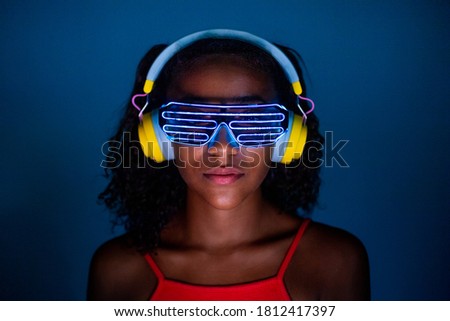 Young woman wearing headphones and futuristic led glasses on blue background - Isolated black woman wearing 3d smart glasses and headphones - virtual reality, future, technology concept  Royalty-Free Stock Photo #1812417397