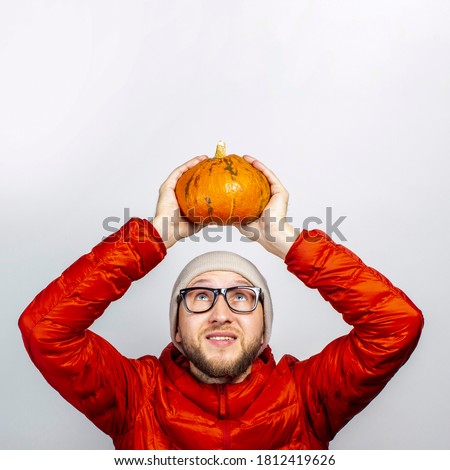 happy young man in a red jacket, hat, holds a pumpkin over his head and looks at it on a light background. Concept of Halloween, celebration, autumn
