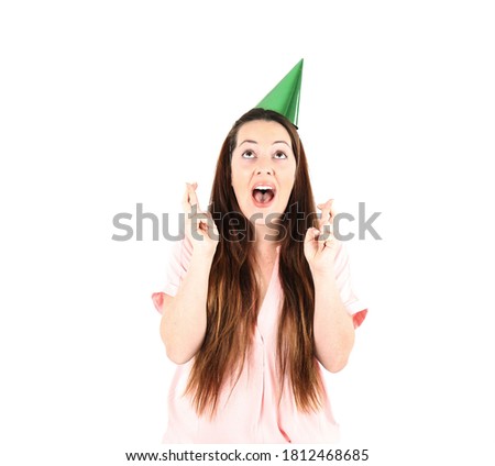 Cute young woman looking up and crossing both her fingers while wearing a green party hat against a white background