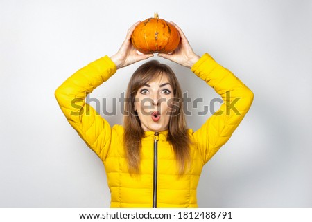 shocked young girl in a yellow jacket holds two pumpkins on a light background. Halloween concept, autumn, celebration
