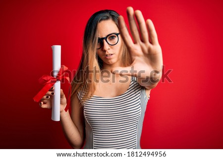 Young blonde woman with blue eyes wearing glasses holding graduated degree diploma with open hand doing stop sign with serious and confident expression, defense gesture