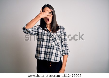 Young brunette woman with blue eyes wearing casual shirt and glasses over white background peeking in shock covering face and eyes with hand, looking through fingers with embarrassed expression.