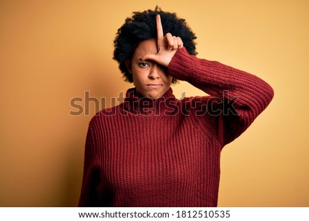 Young beautiful African American afro woman with curly hair wearing casual turtleneck sweater making fun of people with fingers on forehead doing loser gesture mocking and insulting.