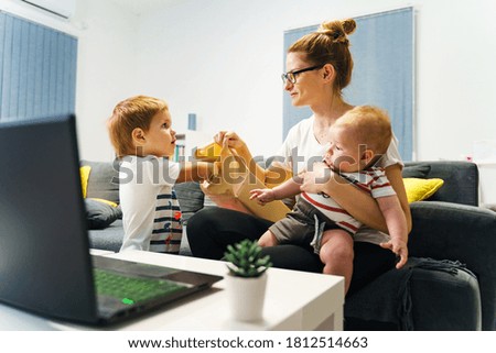 Caucasian single mother hold little baby while helping her son to open birthday present at home in front of the laptop during video call - new normal social distance concept