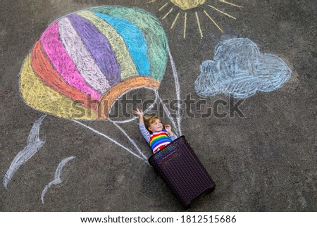 Happy little toddler girl flying in hot air balloon painted with colorful chalks in rainbow colors on ground or asphalt in summer. Cute child having fun. Creative leisure for kids.