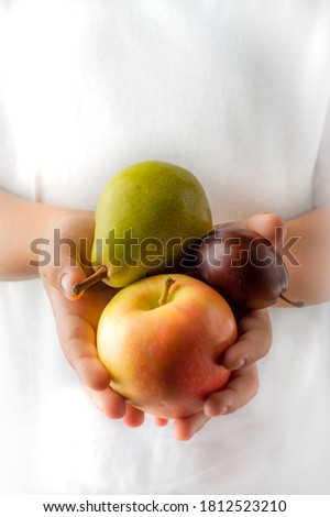 The child holds fruits in his hands: a pear, an apple, a plum, against a background of white clothes.