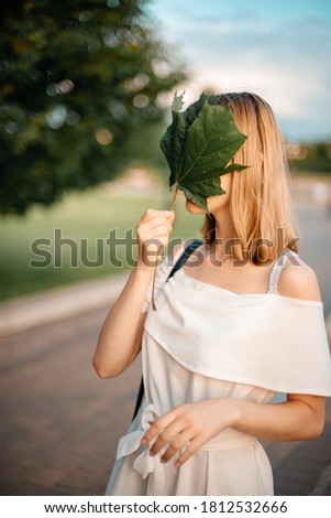 A young attractive girl in a white plain dress covers her face with a green leaf from a maple. Against the background of the forest. Enjoying life. Happiness and fun concept. Copy space.