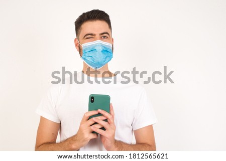Pleased Caucasian man wearing medical mask standing over yellow background, wearing casual clothes using self phone and looking and winking at the camera. Flirt and coquettish concept.