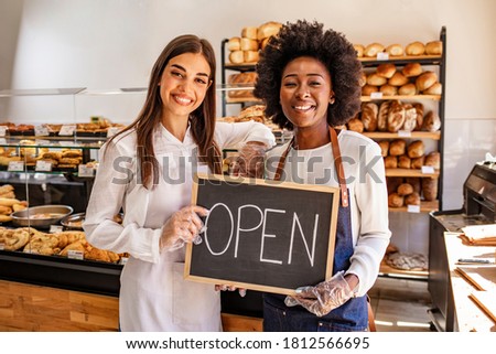 Happy businesswomen standing at bakery shop with open signboard. Close up of women holding sign now we are open support local business. Happy business owner welcoming customers at a bakery shop Royalty-Free Stock Photo #1812566695