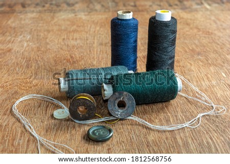 Deep blue, green, black and gray colored sewing thread spools with needle, measuring tape and sewing accessories on brown wooden background.