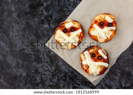 Mini pizza as mummy for kids with cheese, olives and ketchup. Funny crazy Halloween food for children on dark background with copyspace