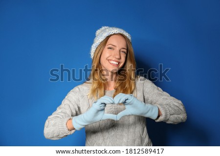 Happy young woman in warm sweater making heart with hands on blue background