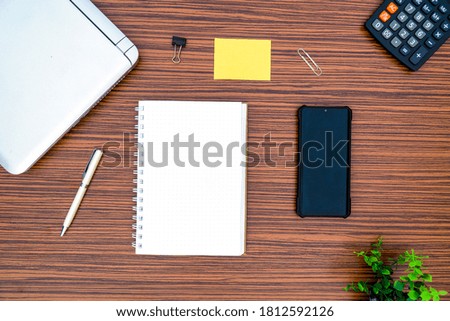 An office table working enviroment. Notepad, sticky note, pen plant, calculator and a lap top on a brown striped zebrawood design table top. Must have objects while working from home during Covid-19