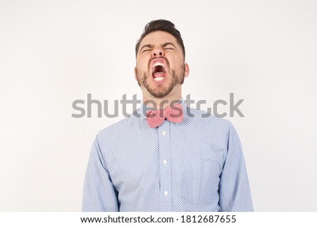 European man dressed in formal shirt and bow tie poses against white studio background angry and mad screaming frustrated and furious, shouting with anger. Rage and aggressive concept.