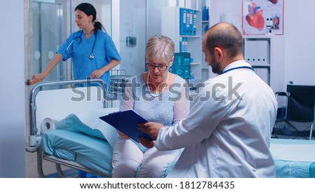 Senior retired woman signing for medical release in modern private clinic sitting on hospital bed, nurse working in background. Healthcare medicine consultation doctor patient