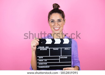 Young beautiful woman wearing glasses over isolated pink background holding clapperboard very happy