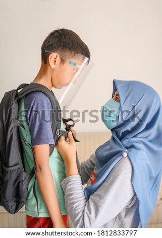 An Asian Indonesian muslim hijab mother preparing her son before going out of home for school during pandemic corona. They use medical masker and face shield to prevent spreading of the virus.  Royalty-Free Stock Photo #1812833797