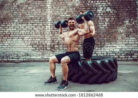 Two muscular athletes training, one raise, push the dumbbell when other is motivating. Scream. Working hard. Street gym. Exercise for the shoulder muscles, deltoid.
