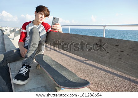 Attractive teenager boy relaxing with a skateboard and sitting down on a bench by the sea, holding a smartphone mobile technology during a sunny day, outdoors. Teens lifestyle.