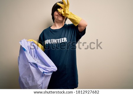 Young down syndrome volunteer woman picking rubbish and plastic from the trash with happy face smiling doing ok sign with hand on eye looking through fingers