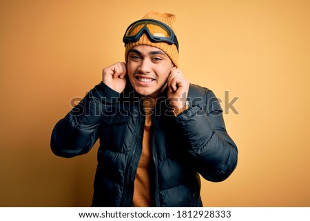 Young brazilian skier man wearing snow sportswear and ski goggles over yellow background Smiling pulling ears with fingers, funny gesture. Audition problem