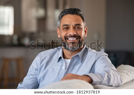 Portrait of happy mid adult man sitting on sofa at home. Handsome latin man in casual relaxing on couch and smiling. Cheerful indian guy looking at camera.  Royalty-Free Stock Photo #1812937819