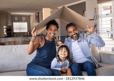 Portrait of smiling family sitting on couch holding cardboard roof and looking at camera. African and indian parents with daughter holding cardboard roof over heads while sitting on sofa in new home  Royalty-Free Stock Photo #1812937846