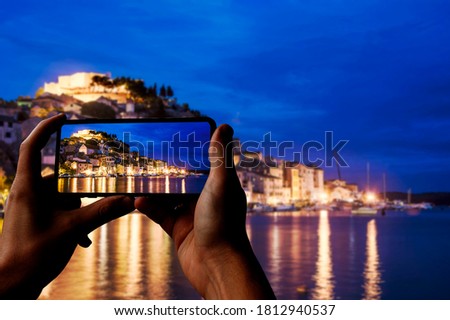 Tourist taking photo of illuminated Sibenik with fortress, old buildings, sea and blue sky. Harbor of Sibenik, Croatia at night, view at blue hour on St. Michael's Fortress