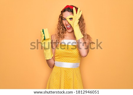 Young beautiful blonde woman with blue eyes cleaning using gloves and cleaner scourer with happy face smiling doing ok sign with hand on eye looking through fingers