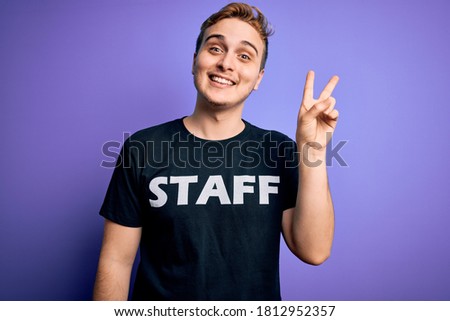 Young handsome redhead worker man wearing staff t-shirt uniform over purple background smiling looking to the camera showing fingers doing victory sign. Number two.
