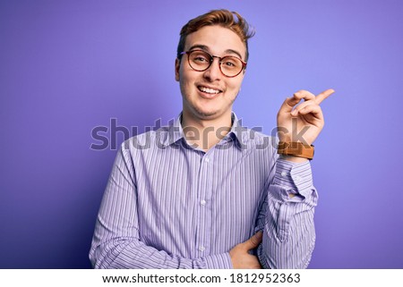 Young handsome redhead man wearing casual shirt and glasses over purple background with a big smile on face, pointing with hand and finger to the side looking at the camera.