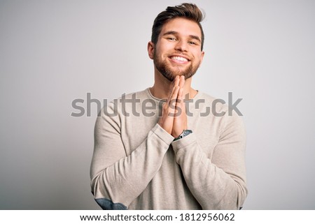 Young handsome blond man with beard and blue eyes wearing casual sweater praying with hands together asking for forgiveness smiling confident.