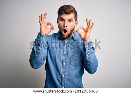 Young handsome blond man with beard and blue eyes wearing casual denim shirt looking surprised and shocked doing ok approval symbol with fingers. Crazy expression