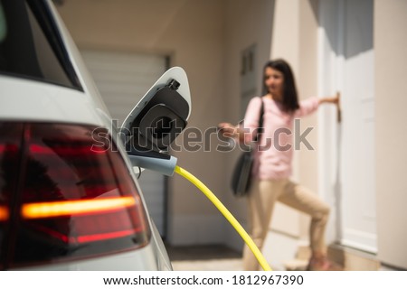 Close up of a electric car charger with female silhouette in the background, entering the home door and locking car Royalty-Free Stock Photo #1812967390