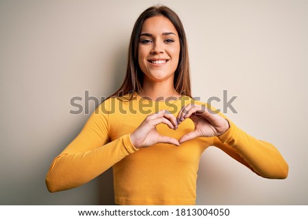Young beautiful brunette woman wearing yellow casual t-shirt over white background smiling in love showing heart symbol and shape with hands. Romantic concept.