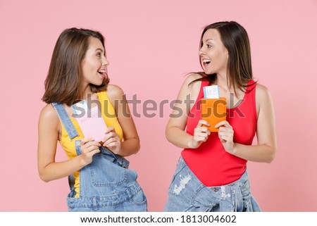 Two surprised young brunette traveler tourist women friends 20s in denim clothes hold passport ticket boarding pass looking at each other isolated on pastel pink colour background studio portrait