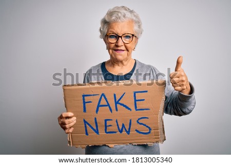 Senior grey-haired woman holding banner with fake news message over white background happy with big smile doing ok sign, thumb up with fingers, excellent sign