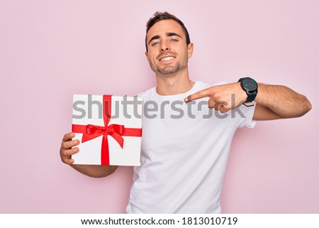 Young handsome man with blue eyes holding birthday present over isolated pink background very happy pointing with hand and finger