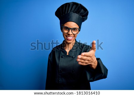 Young african american chef woman wearing cooker uniform and hat over blue background smiling friendly offering handshake as greeting and welcoming. Successful business.