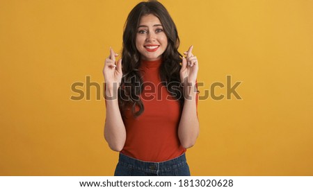 Pretty brunette girl in red top with crossed fingers hopefully praying over colorful background. Please expression