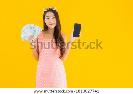 Portrait beautiful young asian woman show a lot of cash and money with mobile smart phone on yellow isolated background