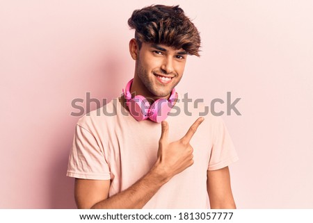 Young hispanic man listening to music using headphones smiling cheerful pointing with hand and finger up to the side 