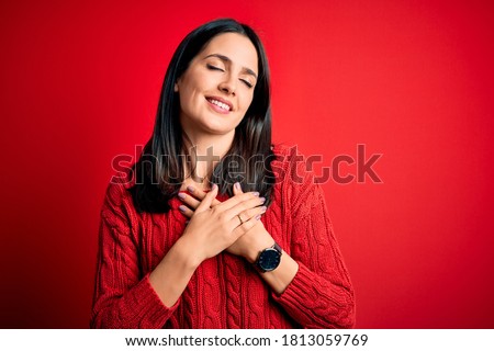 Young brunette woman with blue eyes wearing casual sweater over isolated red background smiling with hands on chest with closed eyes and grateful gesture on face. Health concept.