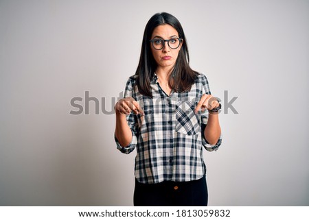 Young brunette woman with blue eyes wearing casual shirt and glasses over white background Pointing down looking sad and upset, indicating direction with fingers, unhappy and depressed.