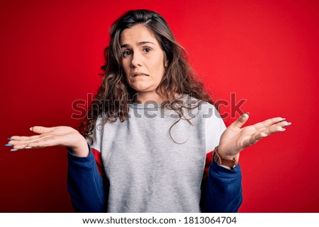 Young beautiful woman with curly hair wearing casual sweatshirt over isolated red background clueless and confused with open arms, no idea concept.