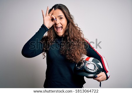 Beautiful motorcyclist woman with curly hair holding moto helmet over white background with happy face smiling doing ok sign with hand on eye looking through fingers