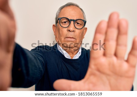 Senior handsome grey-haired man wearing sweater and glasses making selfie by camera with open hand doing stop sign with serious and confident expression, defense gesture