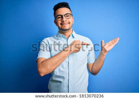 Young handsome man wearing casual summer shirt and glasses over isolated blue background amazed and smiling to the camera while presenting with hand and pointing with finger.